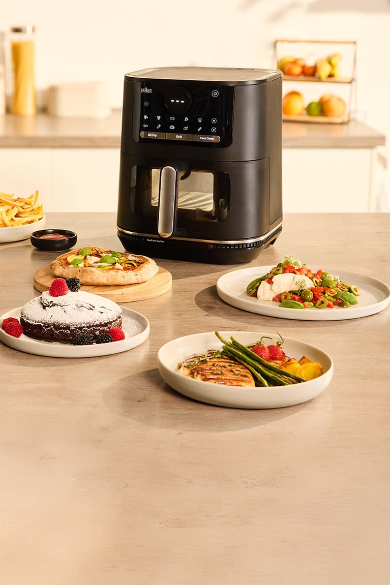 Braun MultiFry 5 Airfryer with Oven, Grill and Airfryer modes standing on a kitchen counter, surronded by plates with chocolate cake, fries, pizza and grilled dishes